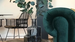A round wooden table with a vase and two glass bottles, a green upholstered chair, and a black metal chair in a modern interior setting. - Hotel La Passerelle (Durbuy)
