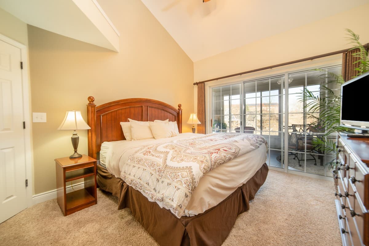A bedroom with a large bed, two bedside tables with lamps, a dresser, and a sliding glass door to a balcony.