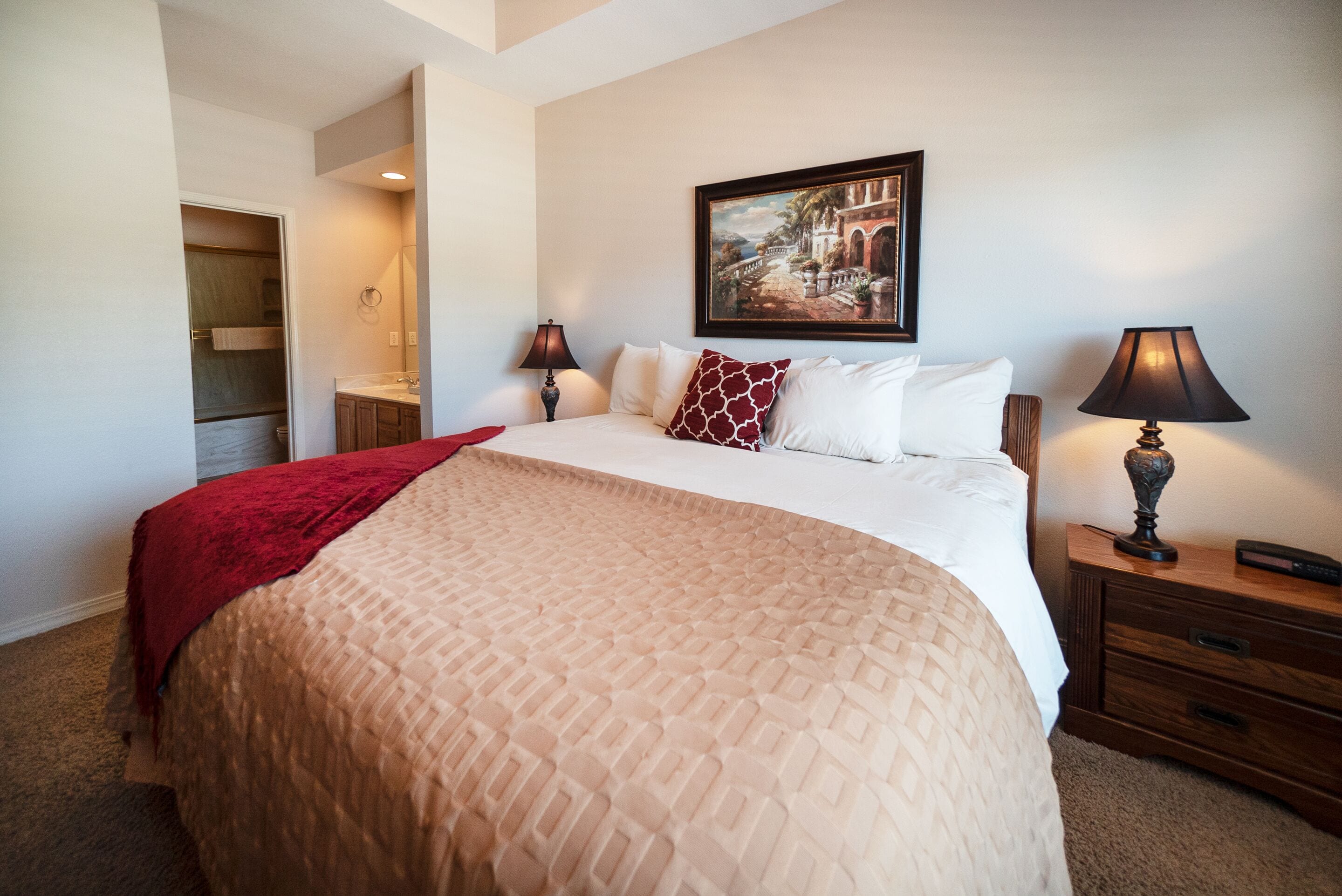 A neatly made bed with a beige quilt, a red throw blanket, and a matching pillow. There is a painting on the wall above the bed, a nightstand with a lamp, and a wooden cabinet with drawers.