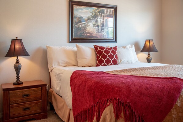 A neatly made bed with a red throw blanket, two bedside lamps, a wooden nightstand with drawers, and a framed picture on the wall.