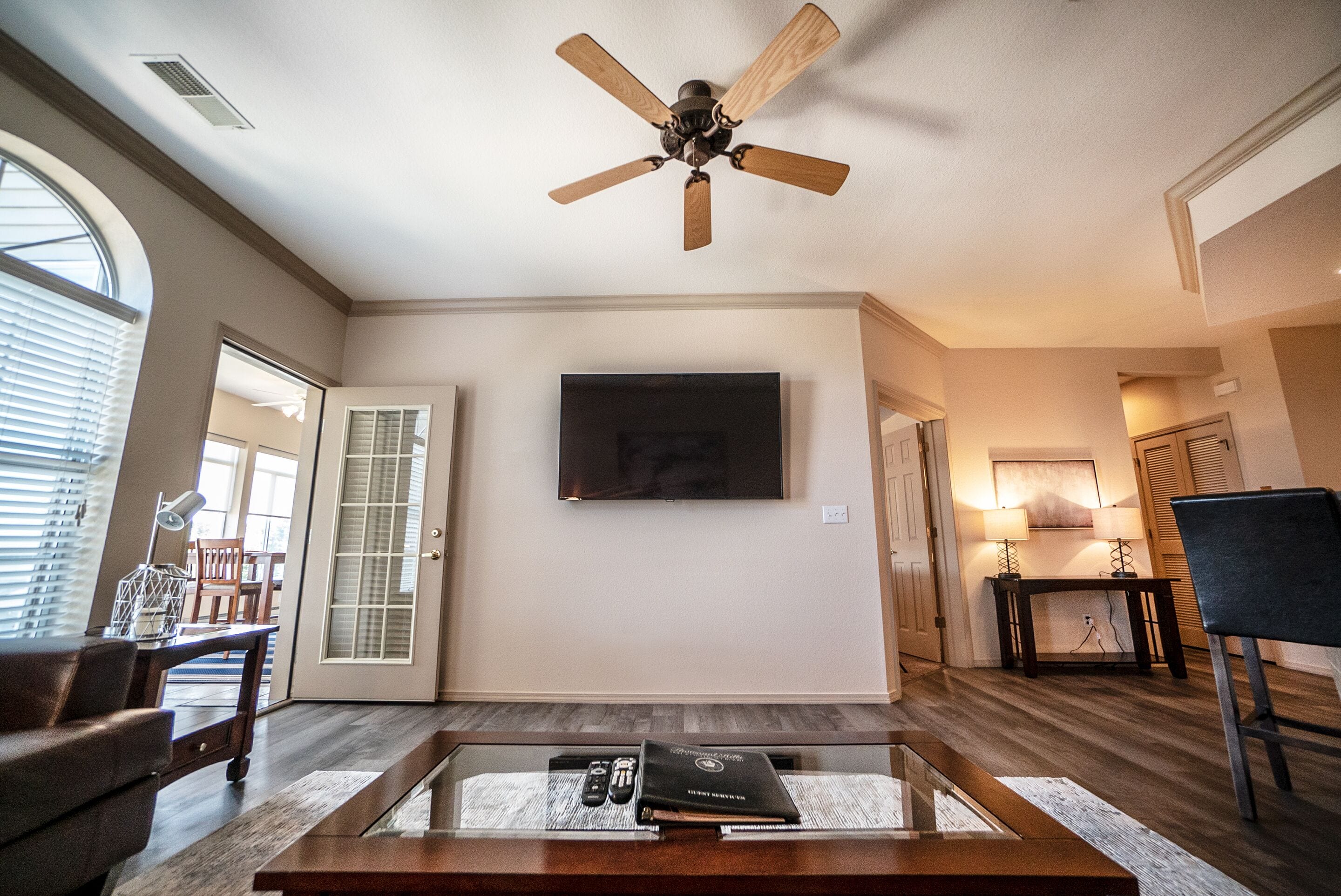 A living room with a ceiling fan, a flat-screen TV, a glass coffee table, and a door leading to another room.