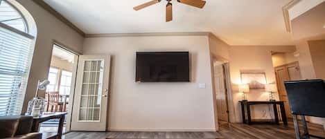 A living room with a ceiling fan, a flat-screen TV, a glass coffee table, and a door leading to another room.