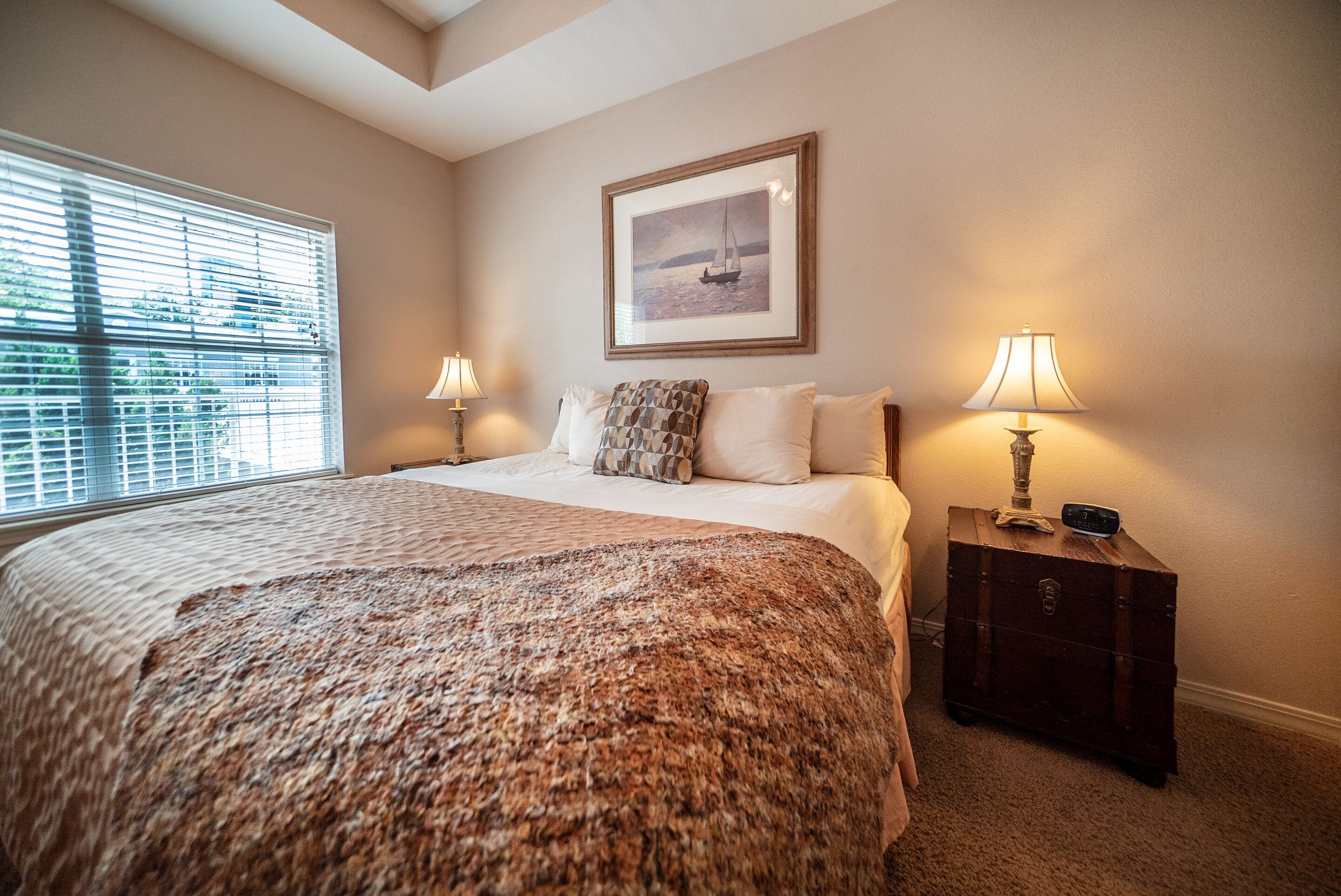 A bedroom with a large bed, two bedside lamps, a framed picture on the wall, and a window with blinds.