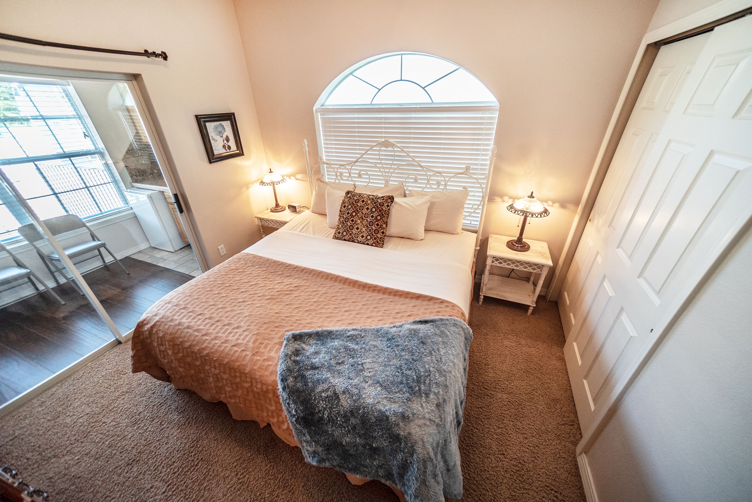 A bedroom with a bed, two bedside tables with lamps, a window with blinds, a door, and a view of a balcony.