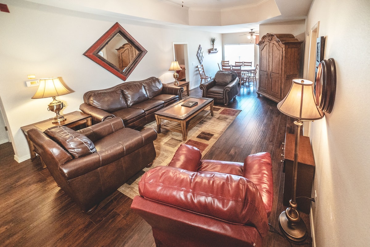 A living room with a brown leather sofa, a red leather armchair, a wooden coffee table, and two lamps. There is a wooden cabinet and a dining table with chairs in the background.