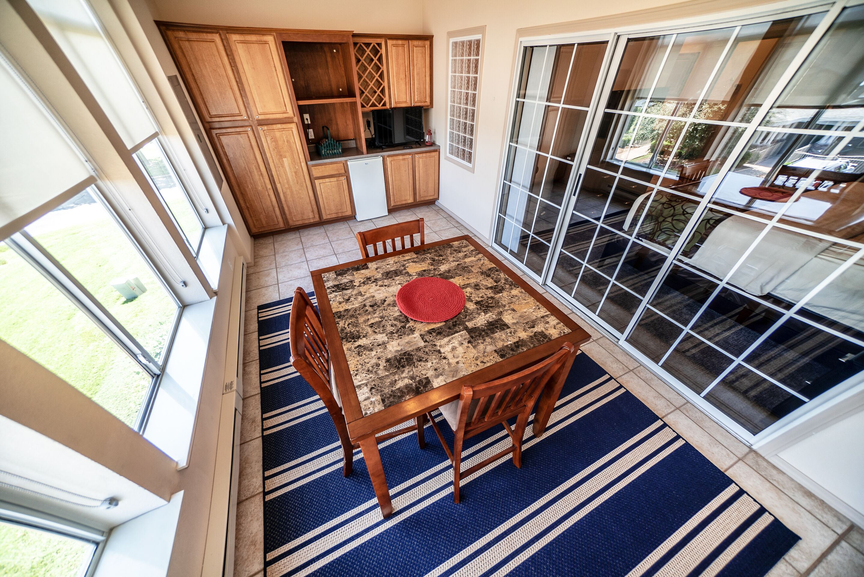 Il y a un coin repas avec une table, des chaises et un set de table rouge. La cuisine est équipée de meubles en bois et d’un réfrigérateur. La pièce est dotée de grandes fenêtres et offre une vue sur l’extérieur.