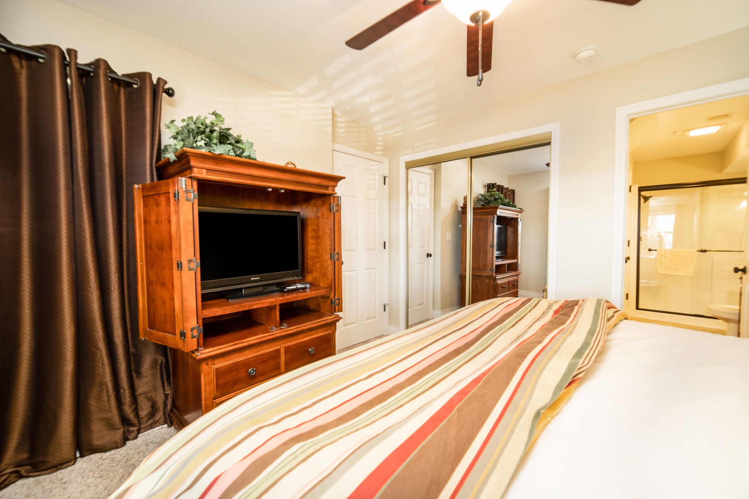 A bedroom with a bed, a television on a wooden cabinet, a ceiling fan, and a door leading to another room.
