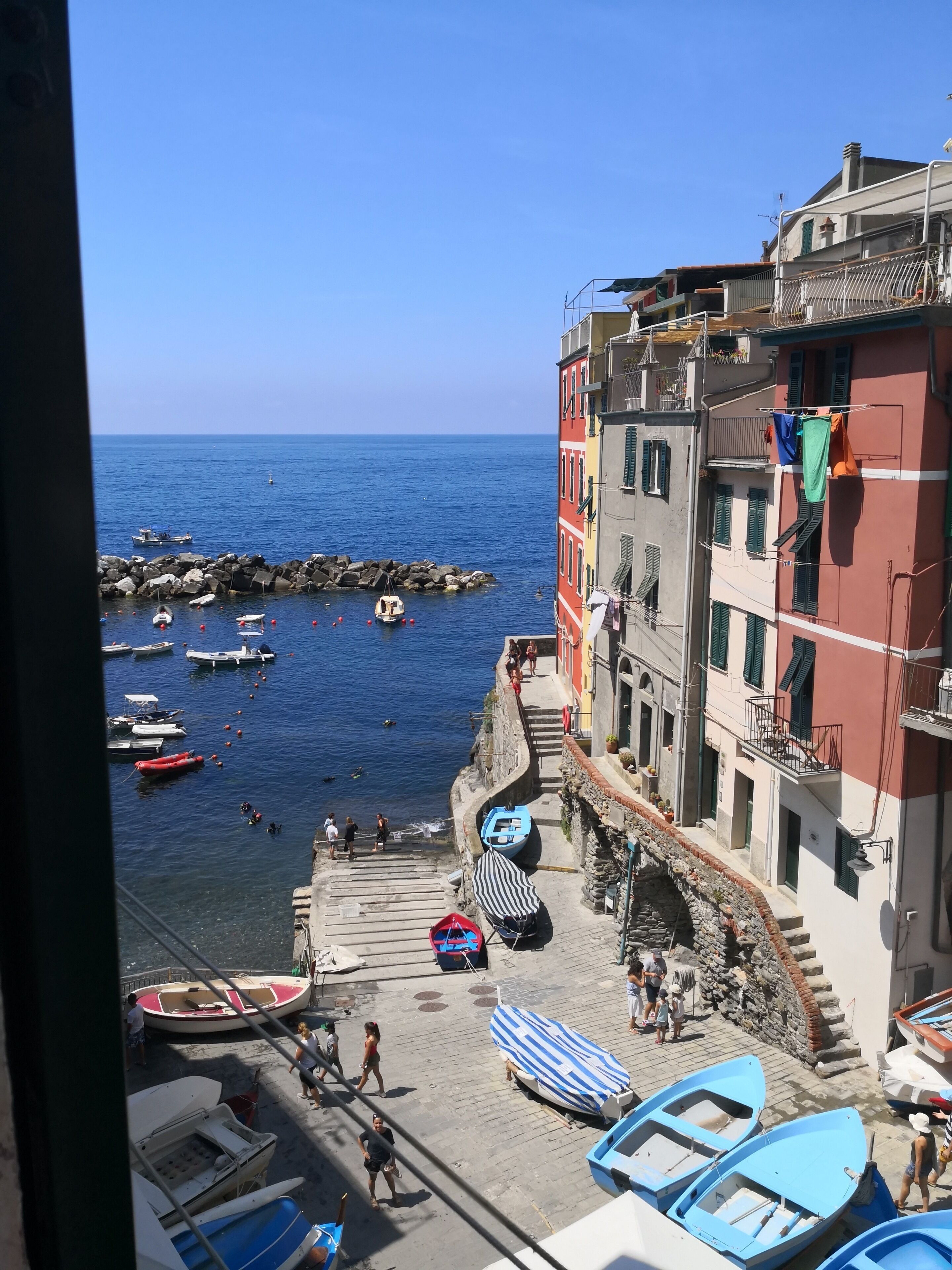 A coastal town with boats docked along the shore, a stone staircase leading to a building, and a clear blue sky.