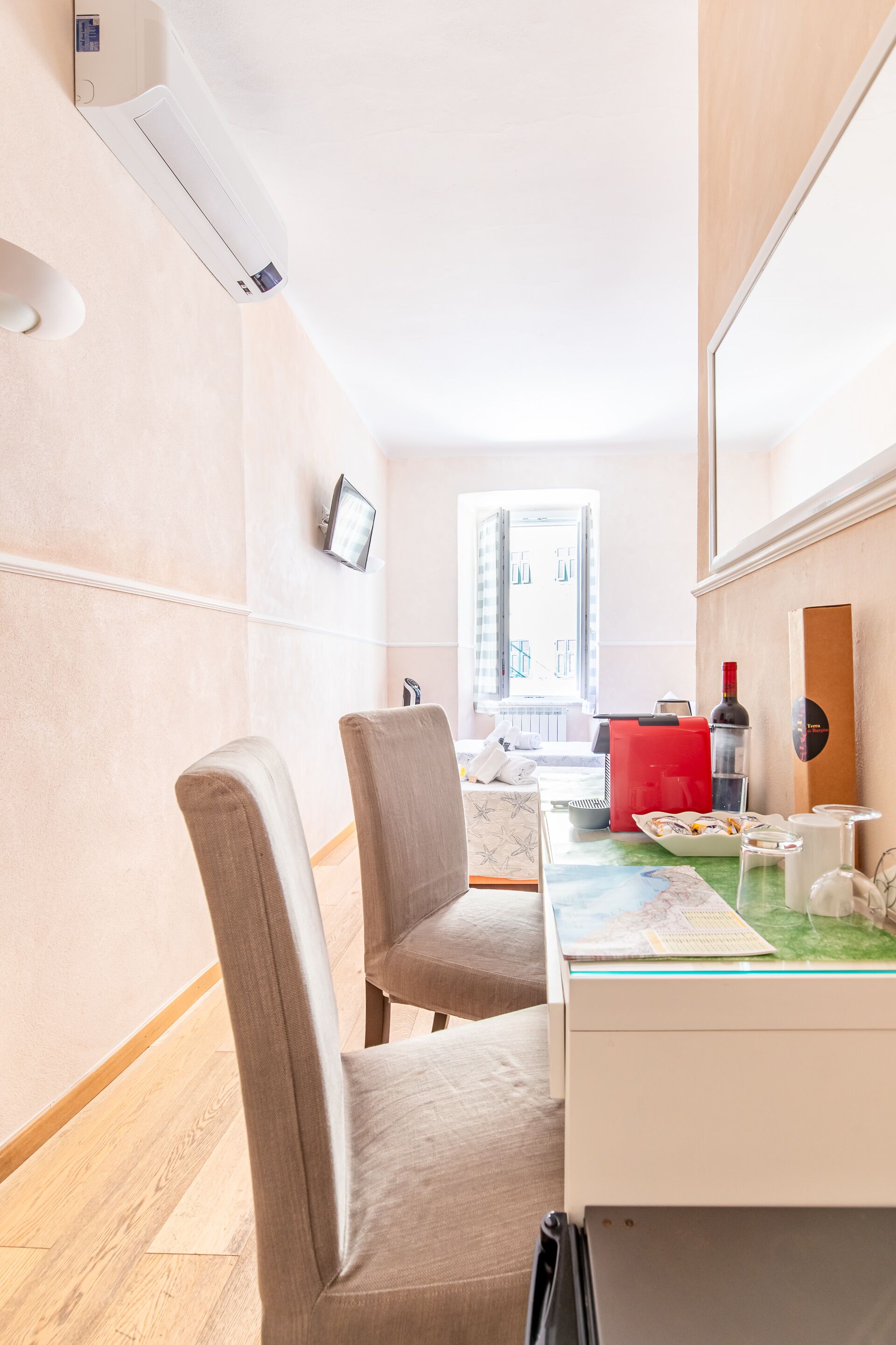 A hotel room with a desk, chairs, a small table with a map, a red suitcase, a wine bottle, and a TV on the wall.