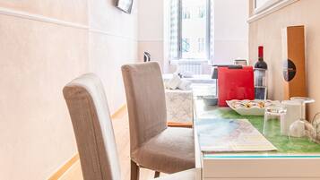 Une chambre d’hôtel avec un bureau, des chaises, une petite table avec une carte, une valise rouge, une bouteille de vin et un téléviseur fixé au mur.