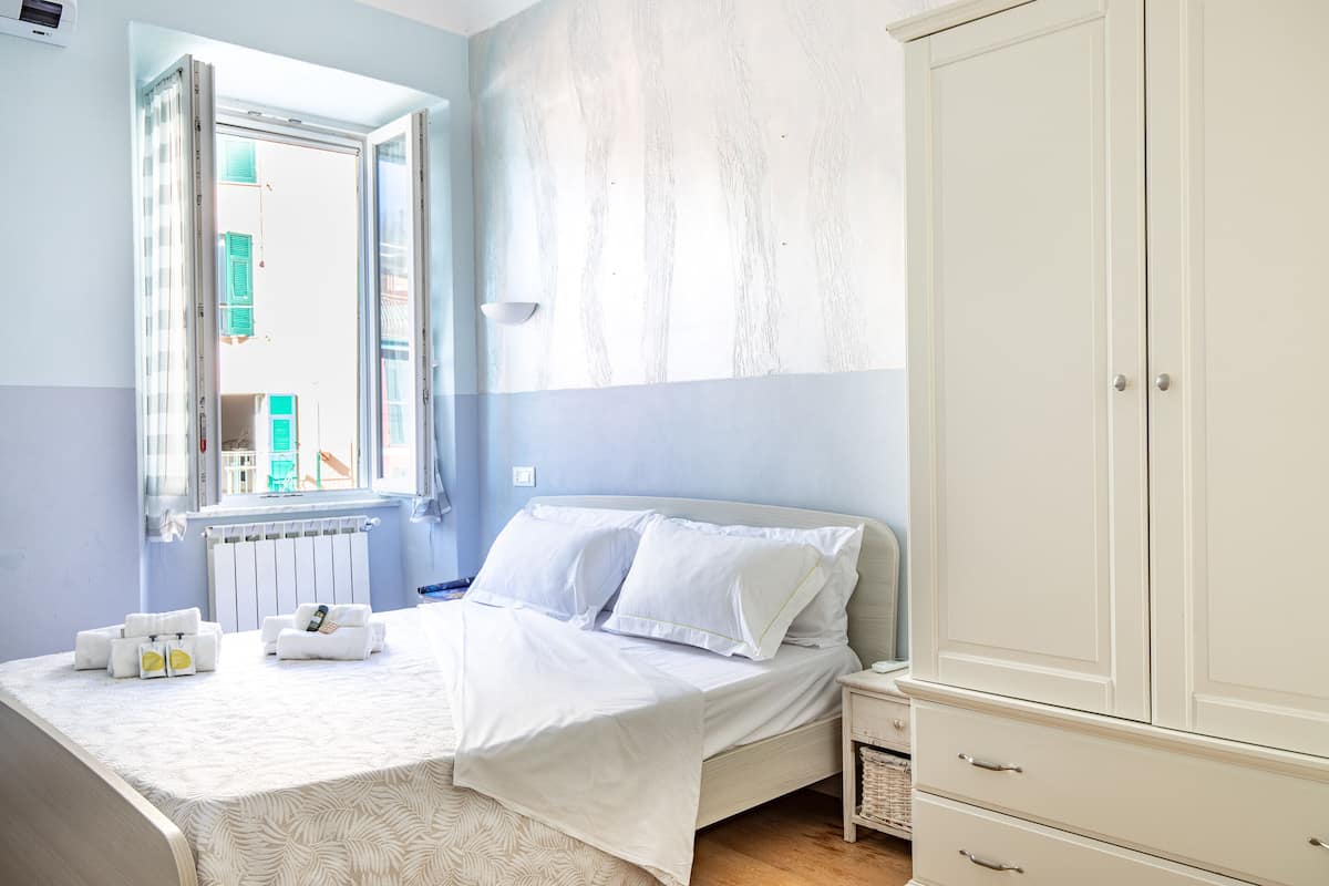 A bedroom with a bed, a wardrobe, a bedside table, and a window with a view of buildings outside.