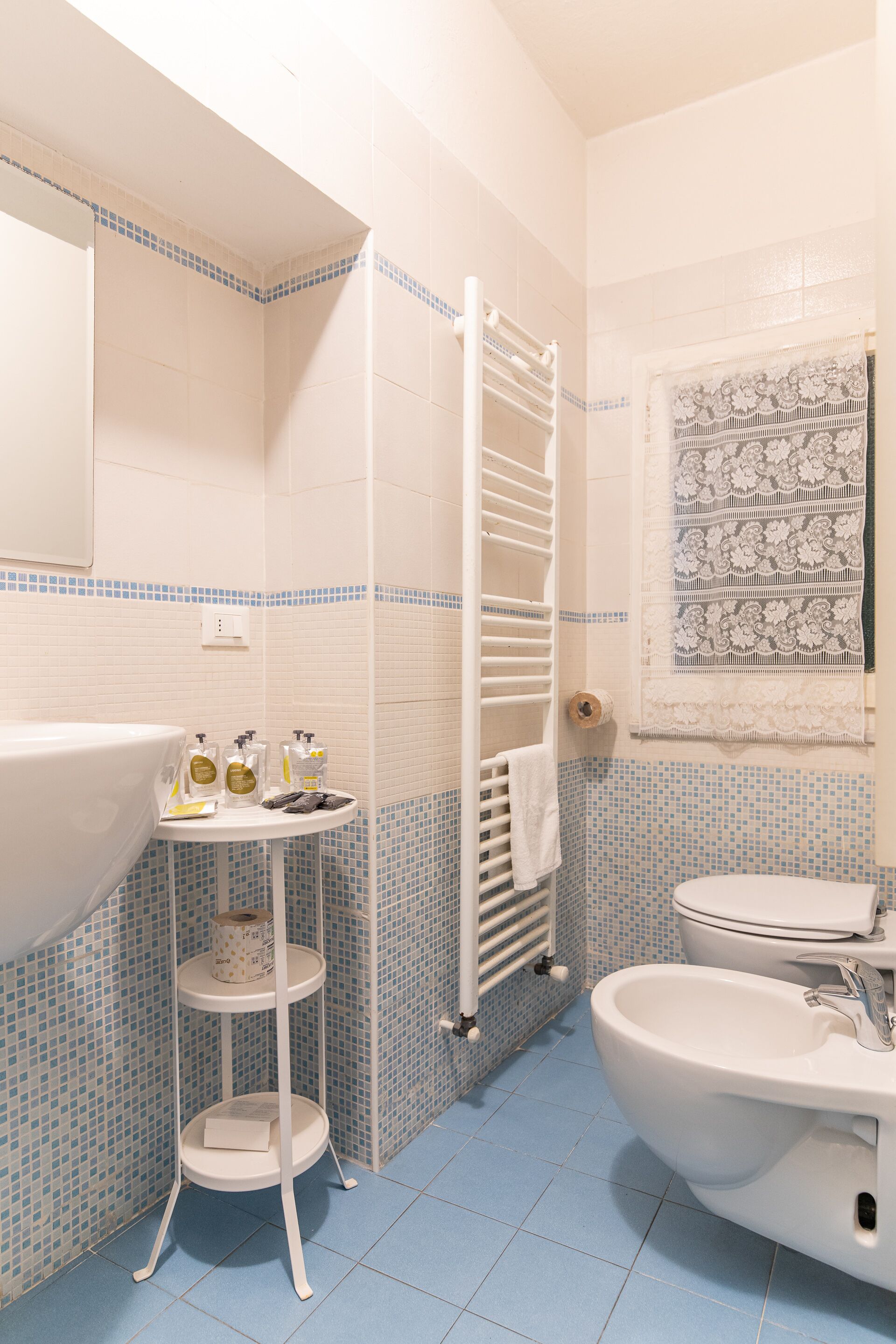 Un bagno con lavandino, WC e bidet, rivestito in piastrelle blu e bianche, con portasci e un piccolo ripiano con articoli da toilette.