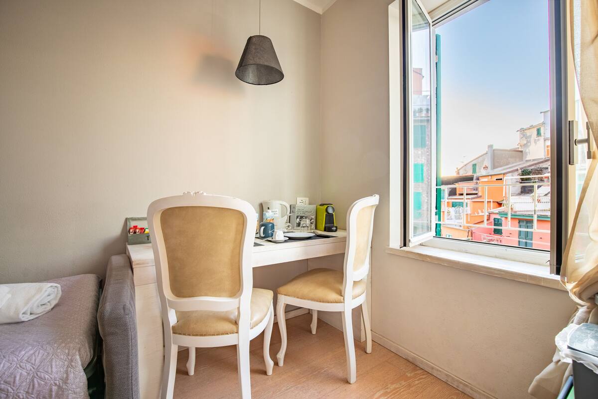 A small, clean room with a bed, a desk, two chairs, a hanging light, and a window with a view of buildings.