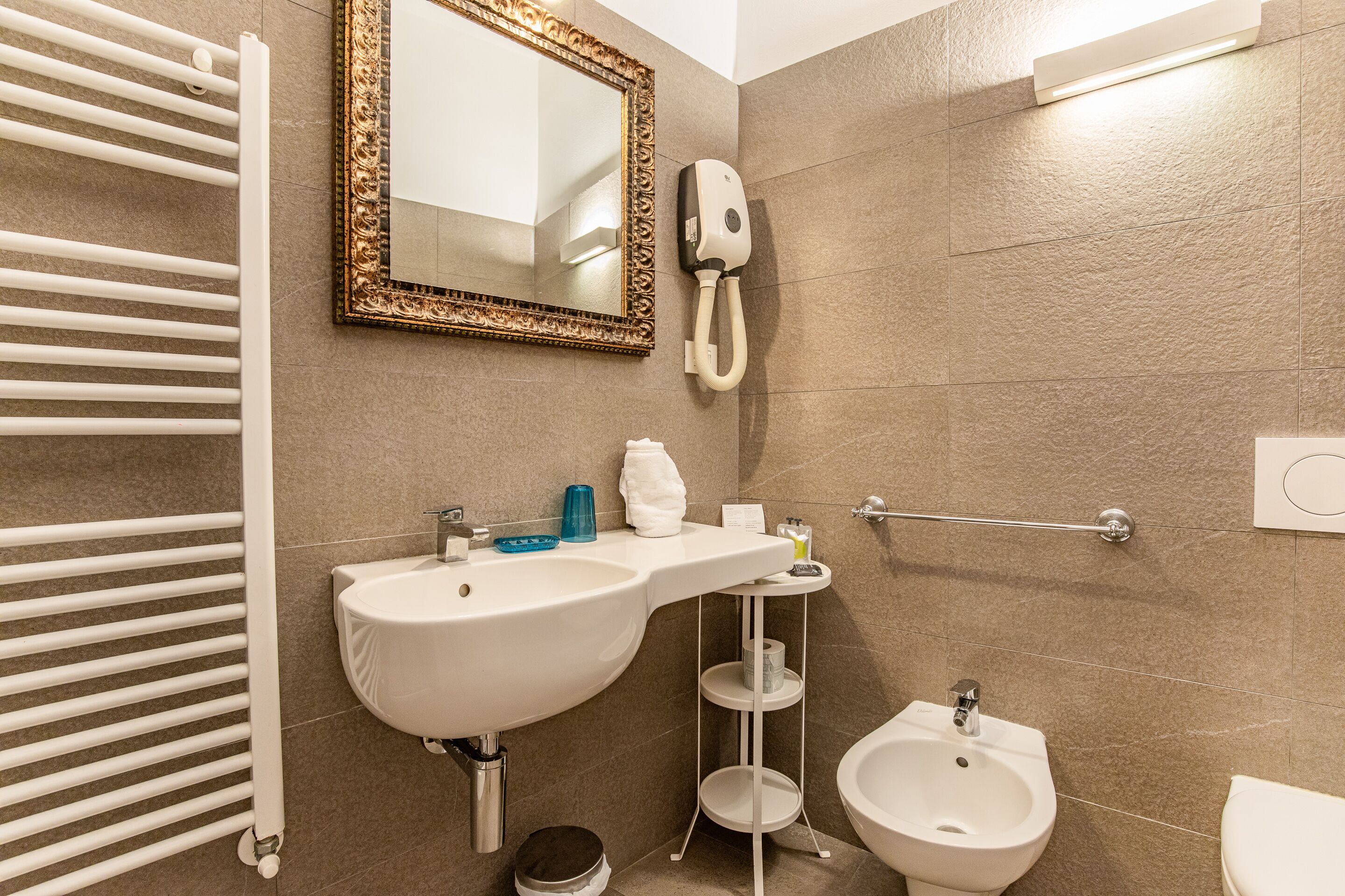A bathroom with a white sink, bidet, and toilet. There is a towel rack, a mirror with an ornate frame, and a soap dispenser.