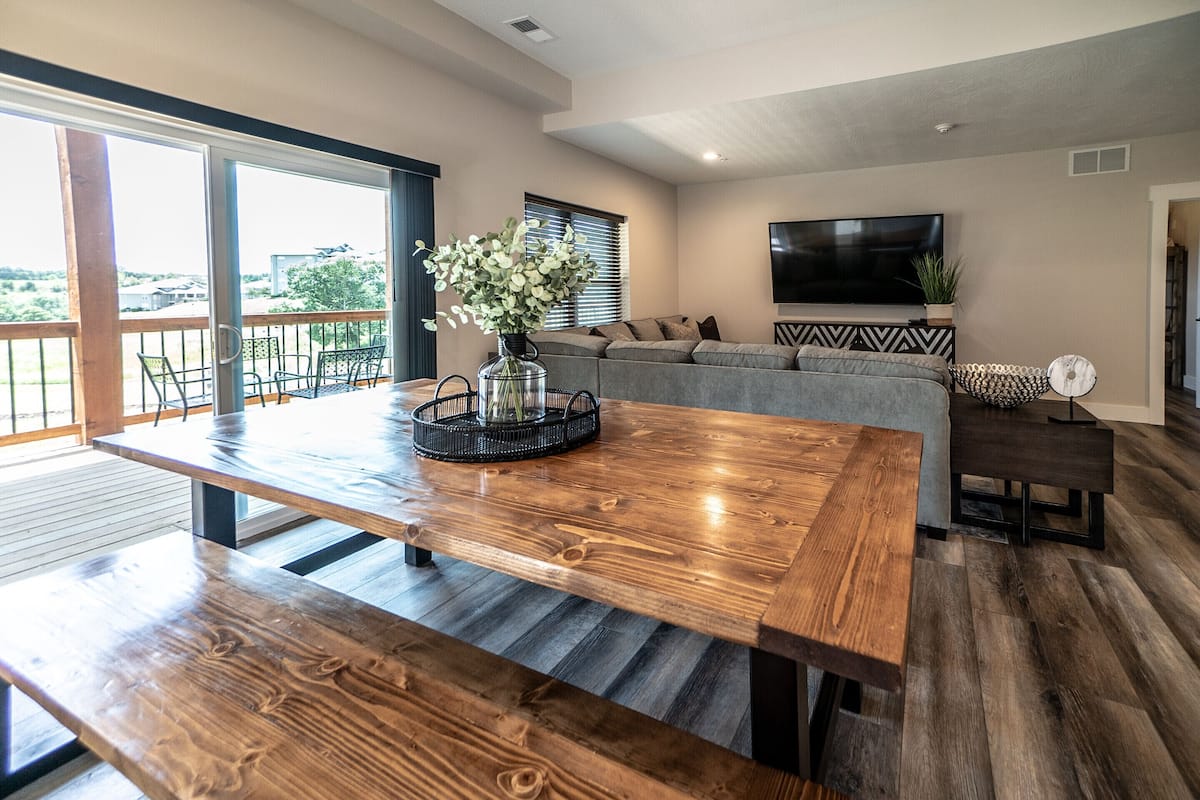 A spacious living room with a large wooden dining table, a grey sofa, and a flat-screen TV.