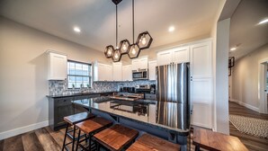 A modern kitchen with white cabinets, a large island with stools, stainless steel appliances, and a hexagonal pendant light. - Reunion Lodges at Thousand Hills (Branson)