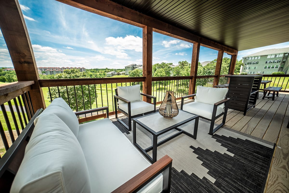 A covered outdoor seating area with white cushioned chairs and tables, a woven floor lamp, and a view of a green landscape and buildings in the distance.