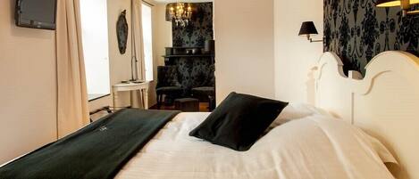 Una cama bien hecha con un almohadón negro y una colcha blanca, un televisor en la pared, un candelabro y un papel tapiz estampado.