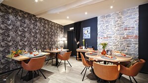 A dining area with two round tables, each surrounded by four orange chairs. The room features a patterned wallpaper on one wall and a stone wall on the other. - Hotel Le Saint Hadelin (Houyet)