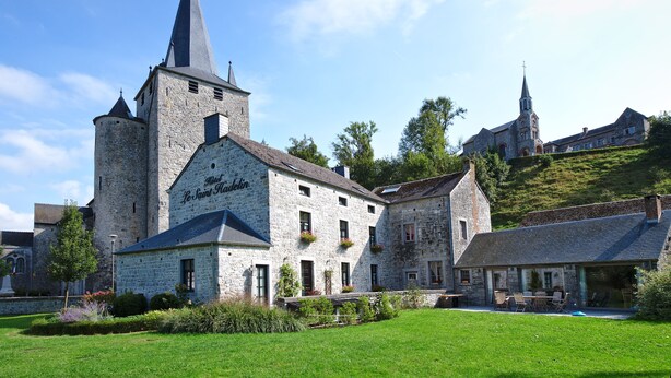 A historic stone building with a church tower, a courtyard with outdoor seating, and a hill with a church in the background. - Hotel Le Saint Hadelin (Houyet)