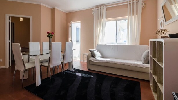 A modern living room with a glass dining table, white chairs, a beige sofa, and a black rug. - Remarkable 2-Bed Apartment (ALVOR, Algarve)