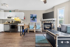 A modern kitchen with white cabinets, a microwave, and a toaster. There is a fireplace with a stone surround and a flat-screen TV mounted above it. A dining area with a wooden table and chairs is visible. - 5 Mins to the Resort & Fernie. On the Elk River (Fernie)