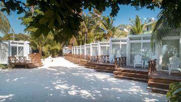 A beachside resort with wooden deck seating and white chairs, surrounded by palm trees and a clear sky.