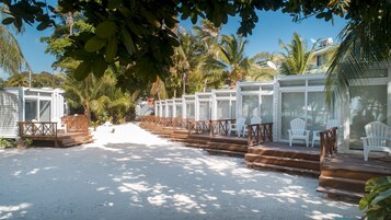 Un resort frente a la playa con sillas blancas y asientos de madera en una terraza, rodeado de palmeras y un cielo despejado.