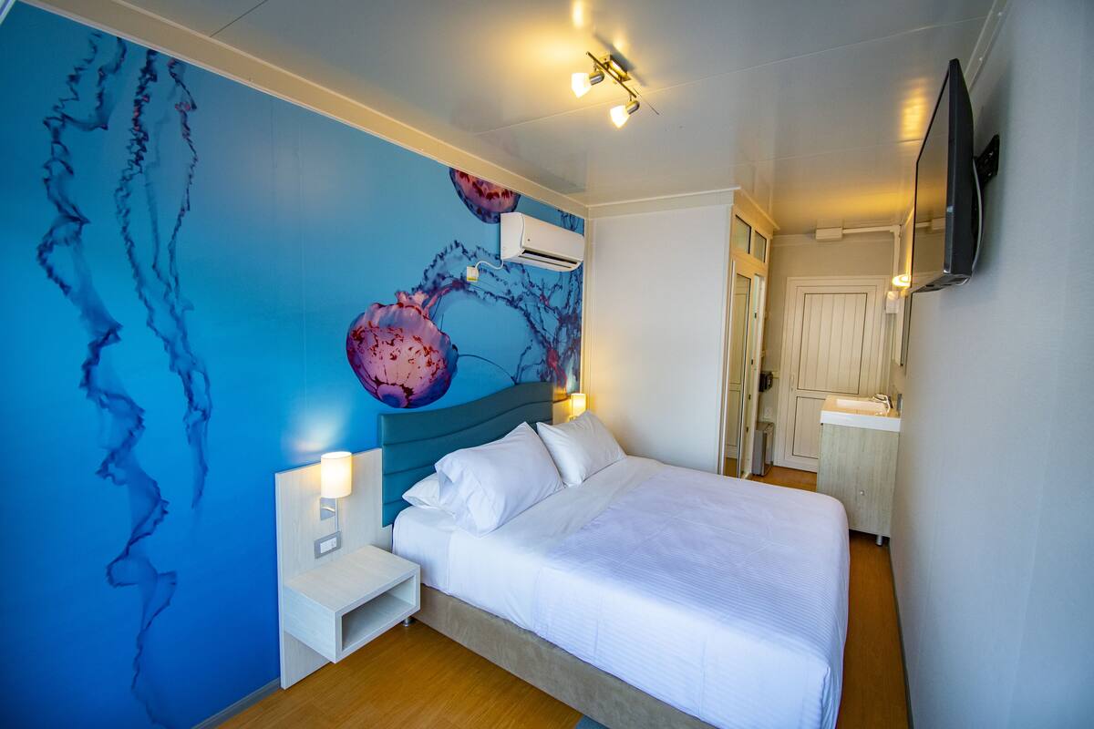 Ein Schlafzimmer mit Bett, Nachttisch, an der Wand montiertem Fernseher, Klimaanlage und einem Badezimmer mit Waschbecken und Spiegel.