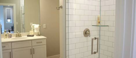 Un baño con ducha revestida de azulejos blancos, cabina de ducha con mampara de vidrio y tocador blanco con espejo.