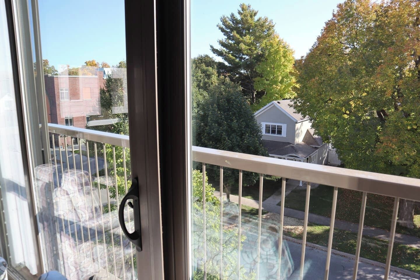 Vista da un balcone con ringhiera in metallo, cielo sereno e zona residenziale con case e alberi.