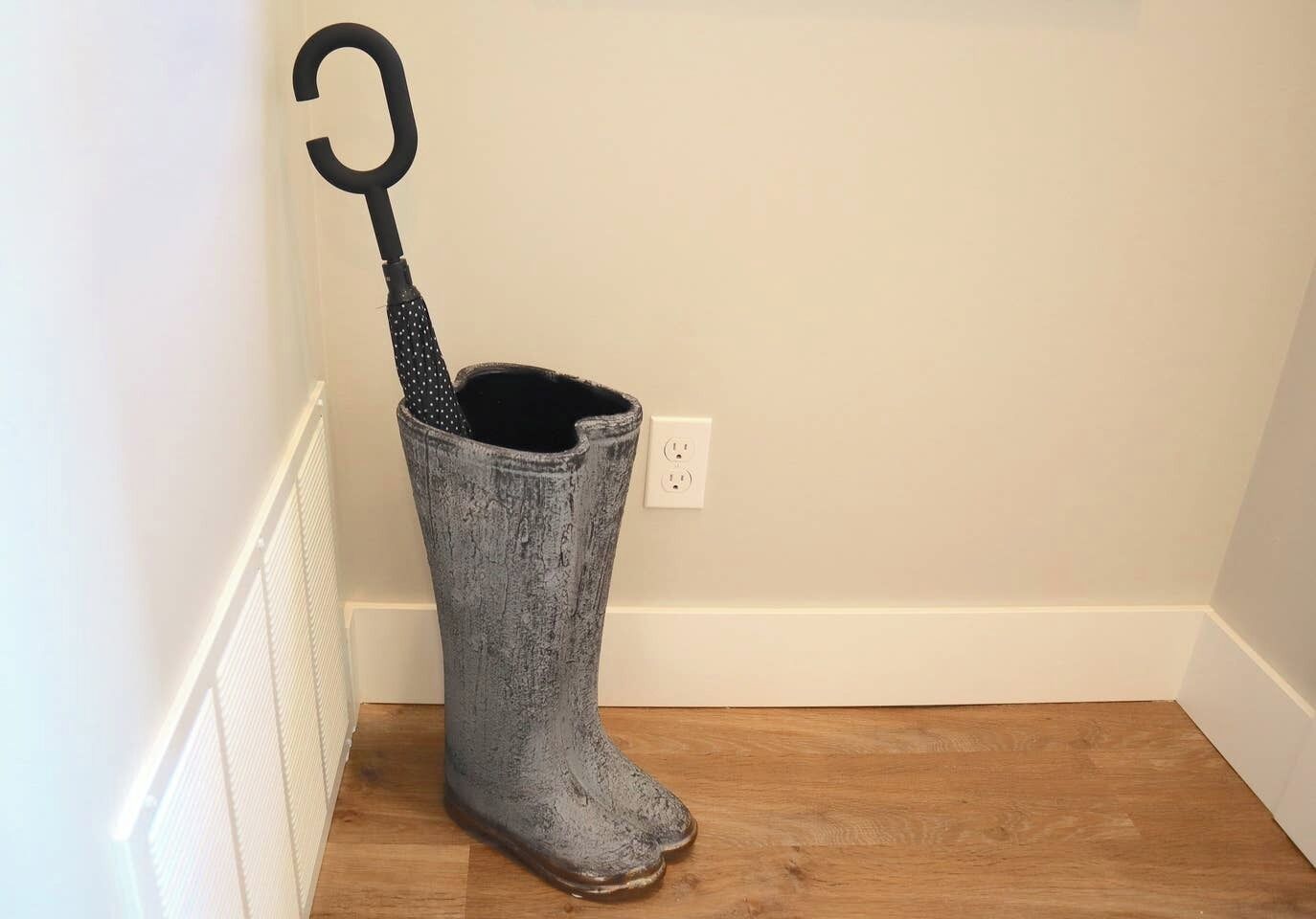 Een grijze regenlaars met een zwarte handgreep op een houten vloer, naast een witte muur met een stopcontact.