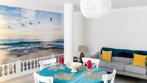 A dining area with a table set for two, a white sofa with yellow cushions, and a large beach mural on the wall. - Laguna Formosa - Holidays in the Algarve (Faro)