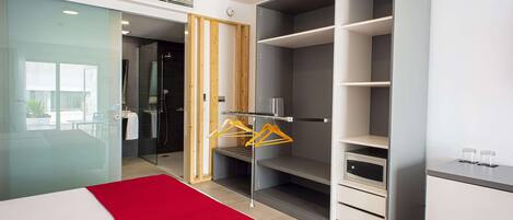 A hotel room with a bed, a red accent wall, a wardrobe, and a bathroom visible through a glass door.