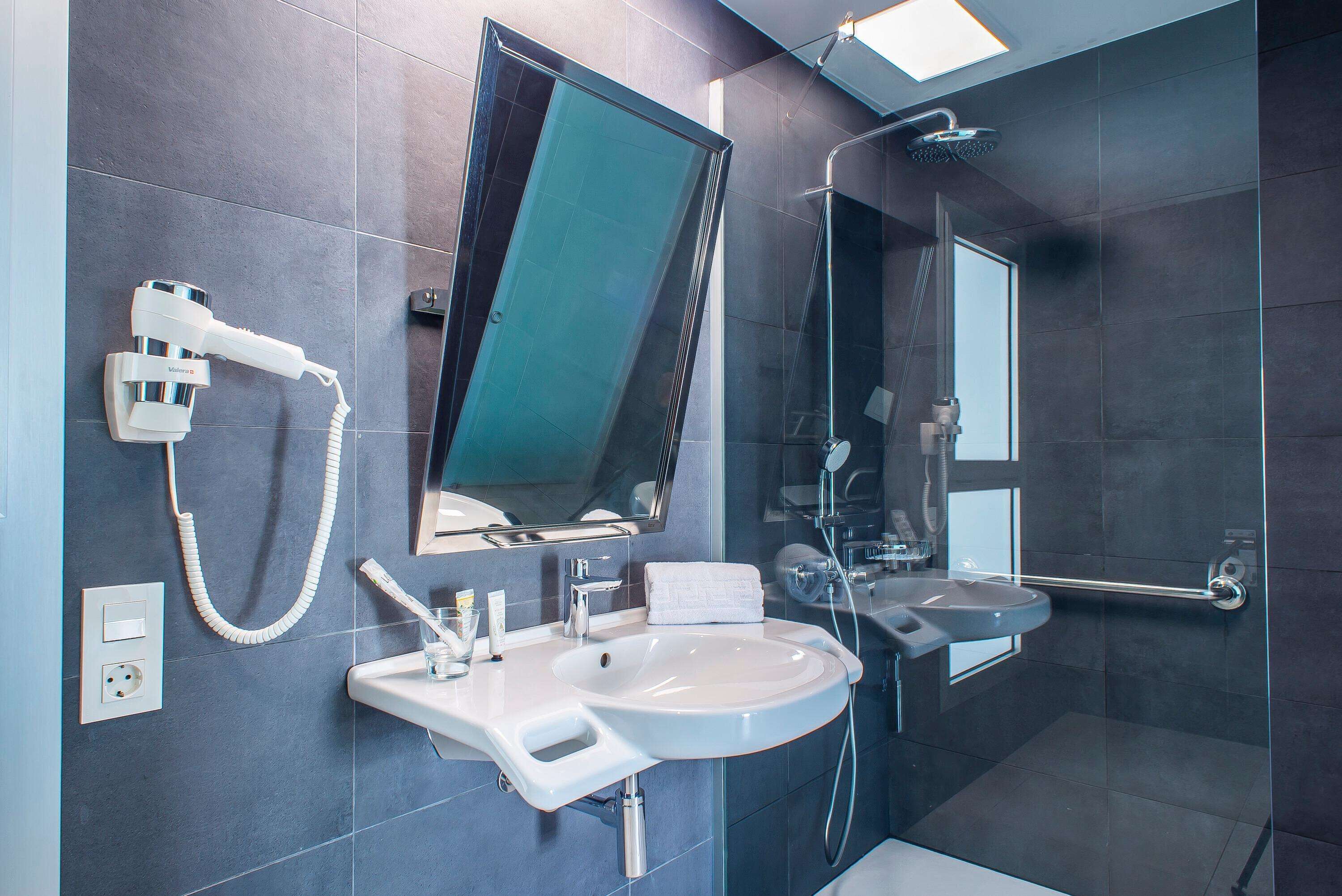 Un baño moderno con un espejo grande, un lavamanos, un cabezal de ducha manual y una ducha con mampara de vidrio.