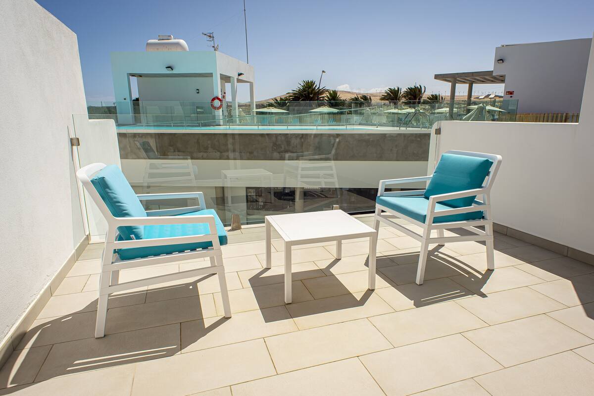 A terrace with two white lounge chairs with blue cushions, a small white table, and a glass balustrade overlooking a pool and distant landscape.
