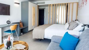 Una habitación de hotel moderna con una cama, un sofá gris, una mesita de madera con una botella y vasos, un televisor y una lámpara de pared.