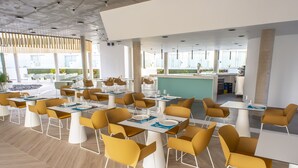 Un comedor moderno con sillas amarillas y mesas blancas, ventanas amplias y una cocina al fondo.