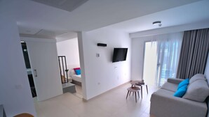 Una sala de estar moderna con un sofá, un televisor montado en la pared, una mesa de comedor y una escalera que conduce a otra habitación.