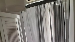 A bedroom with a bed, a window with sheer curtains, a closed door, and a bedside table with a lamp.
