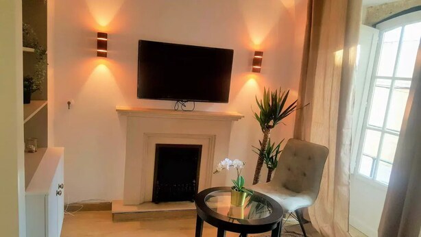 A living room with a flat-screen TV mounted above a fireplace, a comfortable armchair, and a small round table with a vase of flowers.