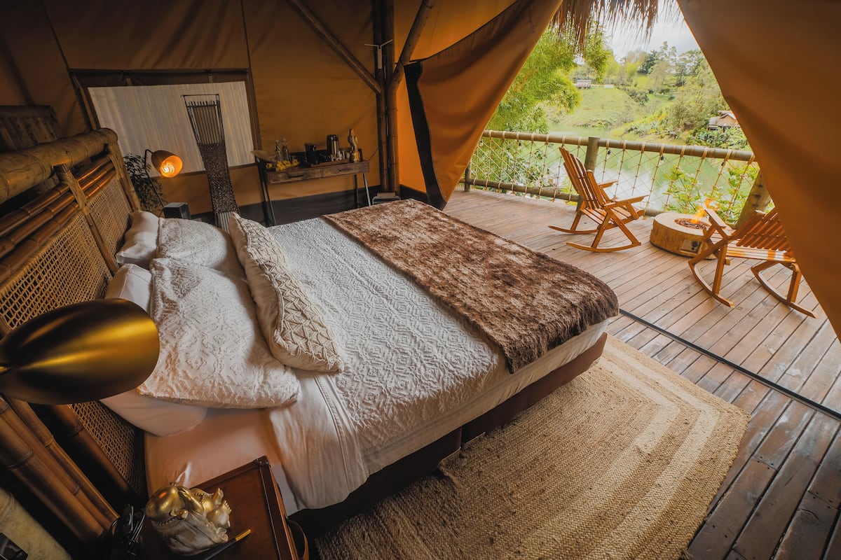 A cozy tented room with a bed, a nightstand, a lamp, and a view of a river and greenery outside.