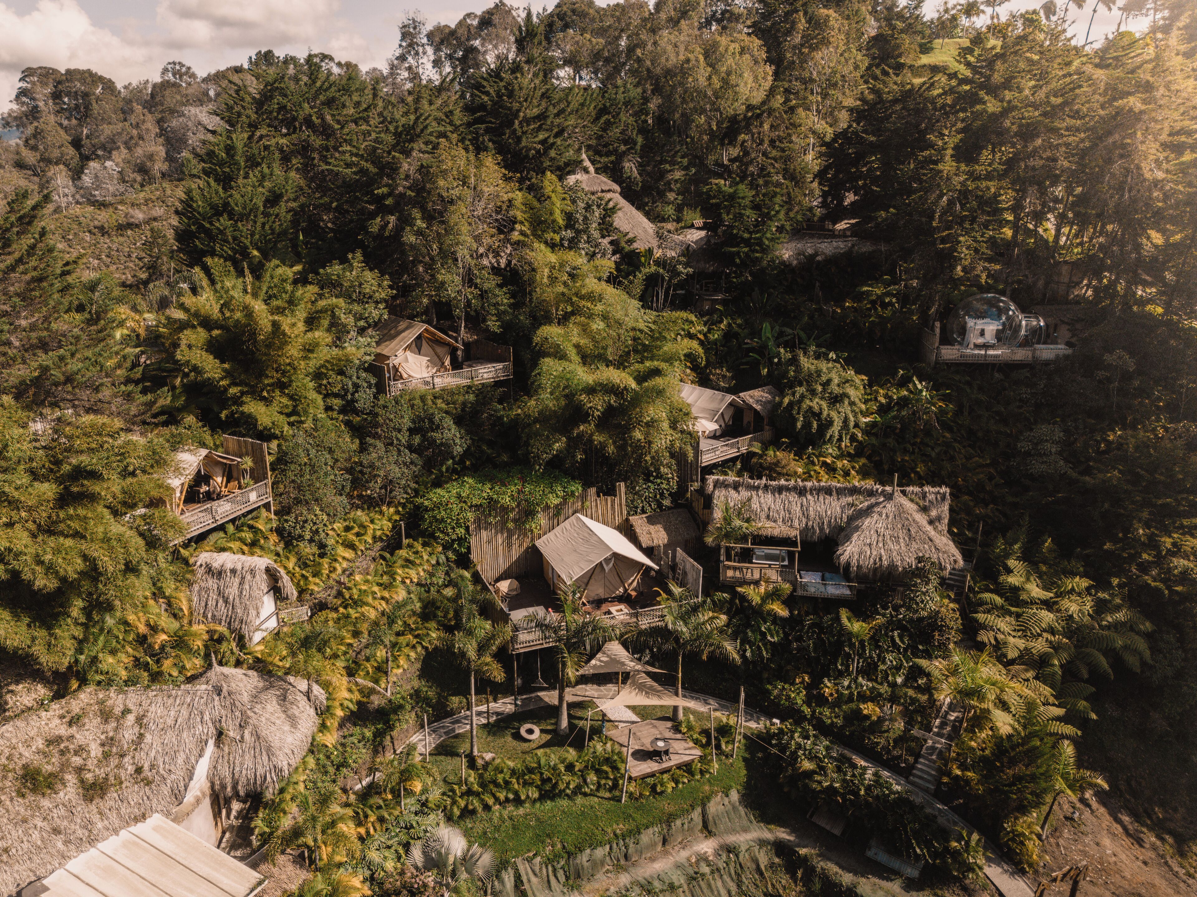 Un chalet écologique isolé, niché au cœur d’une forêt luxuriante, avec des cabanes aux toits de chaume et un espace commun central doté d’une petite piscine.
