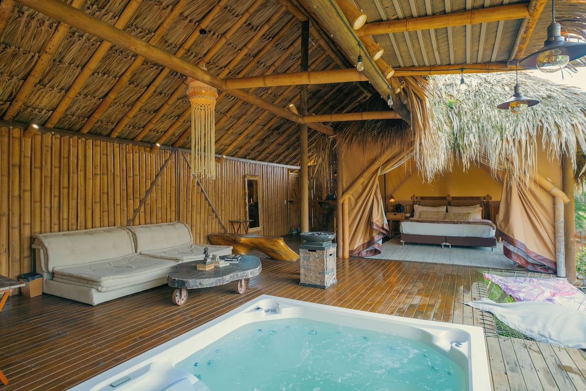A wooden deck with a hot tub, a sofa, and a bed under a thatched roof.