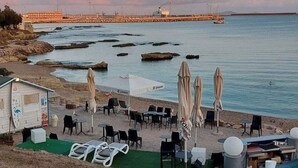 A beachside area with outdoor seating, a small building, and a view of the sea and a pier in the background.