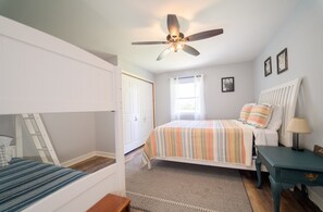 Ein Schlafzimmer mit einem Etagenbett, einem Einzelbett mit farbig gestreifter Bettdecke, einem Deckenventilator, einem Fenster mit Vorhängen und einem kleinen Nachttisch mit Lampe.