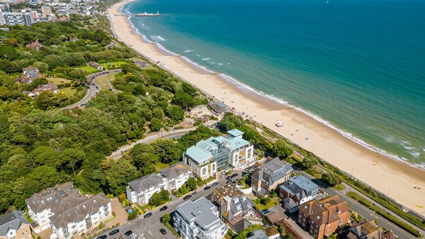 Vista aérea de un pueblo costero con una playa, edificios residenciales y un hotel destacado con techo verde.
