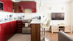 Une cuisine moderne avec des meubles rouges, un îlot central et un four encastré. Un téléviseur est fixé au mur, au-dessus d’un meuble en bois.