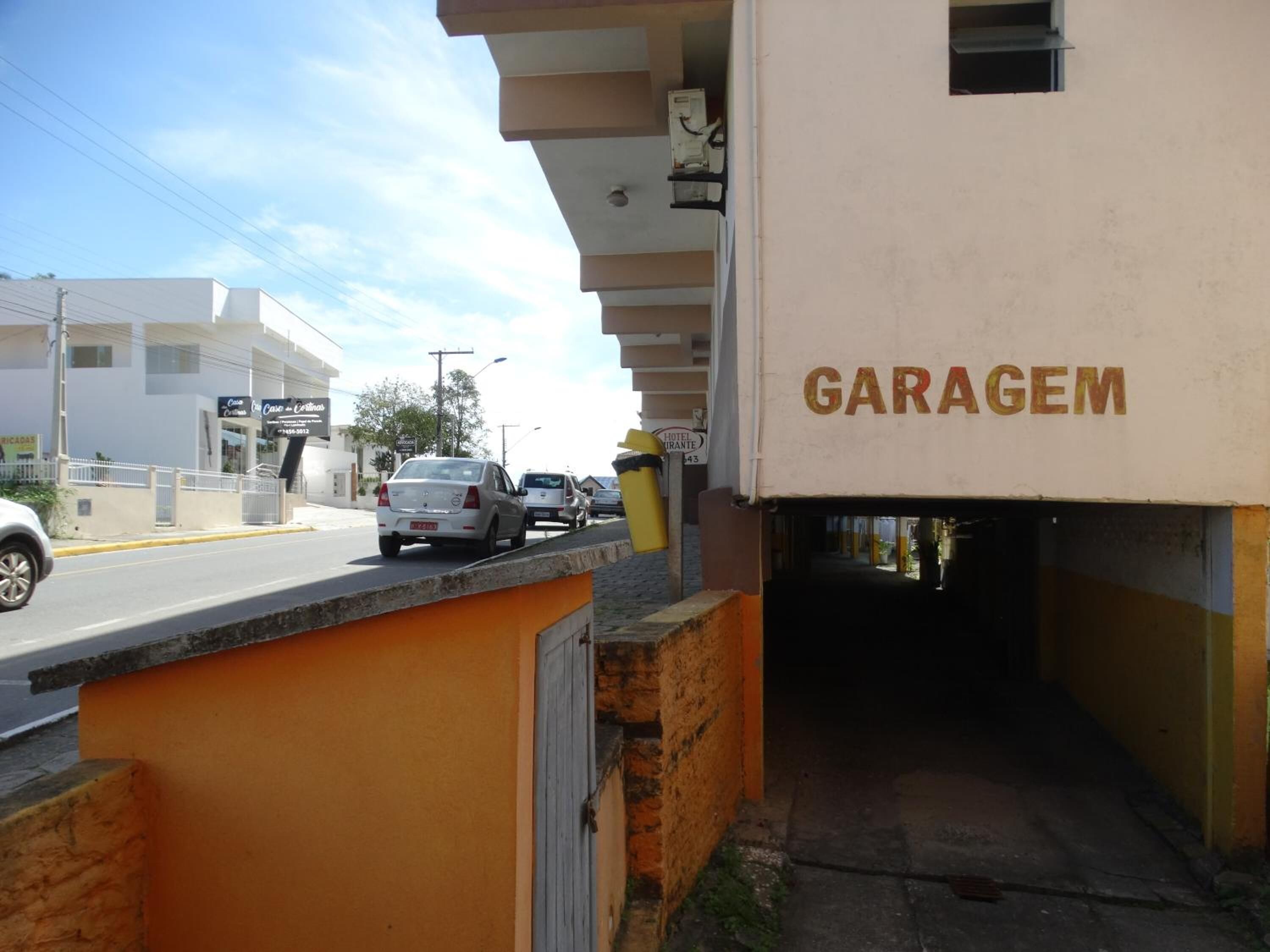 Um edifício com a placa 'GARAGEM', entrada coberta, rua com carros e uma lixeira amarela.