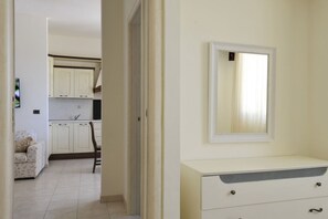 Ein Zimmer mit einem weißen Schminktisch, einem Spiegel und Blick in eine Küche mit weißen Schränken und einem Stuhl.